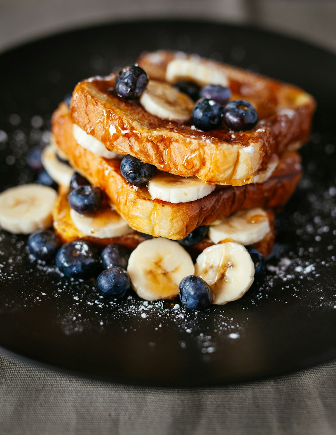 Das Bild zeigt ein appetitliches Gericht aus gestapelten French Toast mit Bananenscheiben und Blaubeeren. Es sieht nach einem süßen Frühstück oder Dessert aus. Die Zutaten sind auf einem dunklen Teller angerichtet und mit Puderzucker bestäubt.